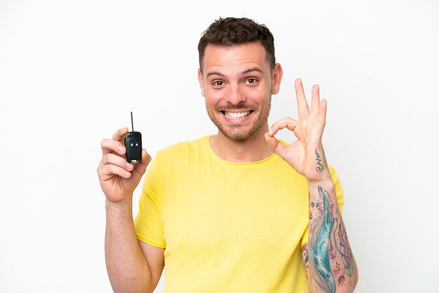 손가락으로 확인 표시를 보여주는 흰색 배경에 고립 된 자동차 키를 들고 젊은 남자