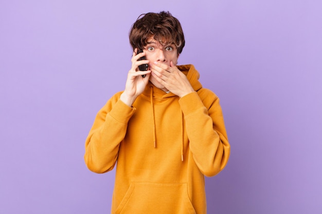 Young man holding a cell covering mouth with hands with a shocked