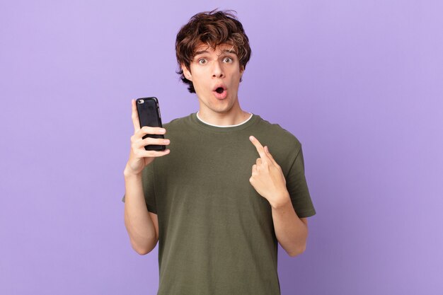 Young man holding a cell looking shocked and surprised with mouth wide open, pointing to self