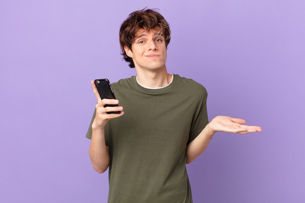 Young man holding a cell shrugging, feeling confused and uncertain