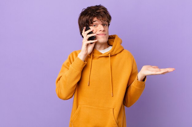 Young man holding a cell shrugging, feeling confused and uncertain