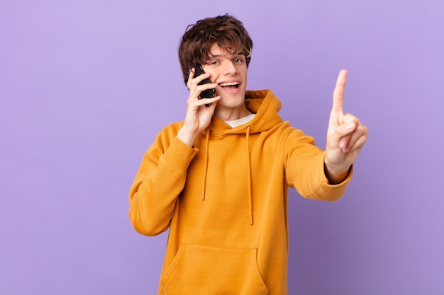 Young man holding a cell smiling and looking friendly, showing number one