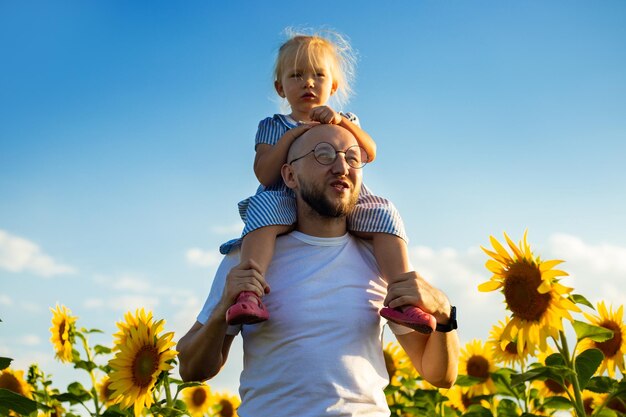 Young man holding a child on his shoulders against a background of blue sky on a sunflower field