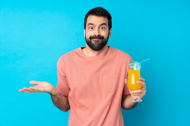 Young man over holding a cocktail over isolated blue wall having doubts while raising hands