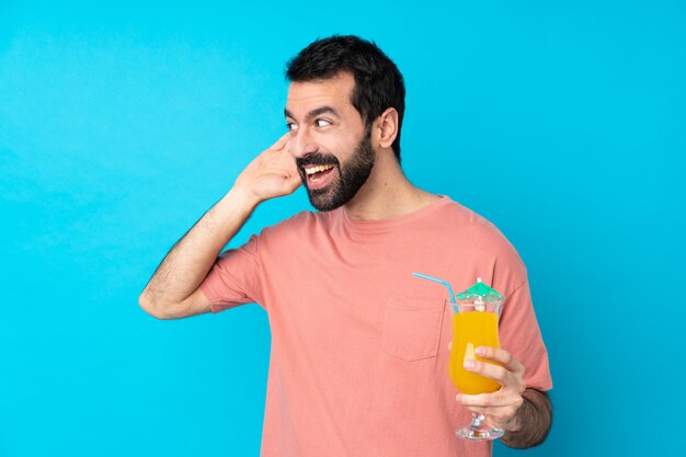 Young man over holding a cocktail over isolated blue wall listening to something by putting hand on the ear