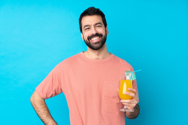 Young man over holding a cocktail over isolated blue wall posing with arms at hip and smiling