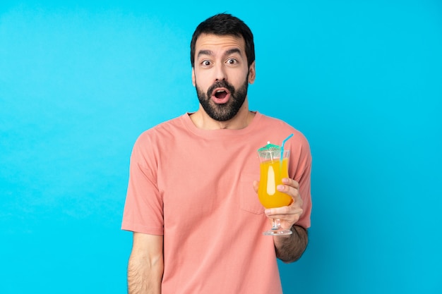 Young man over holding a cocktail over isolated blue wall with surprise facial expression