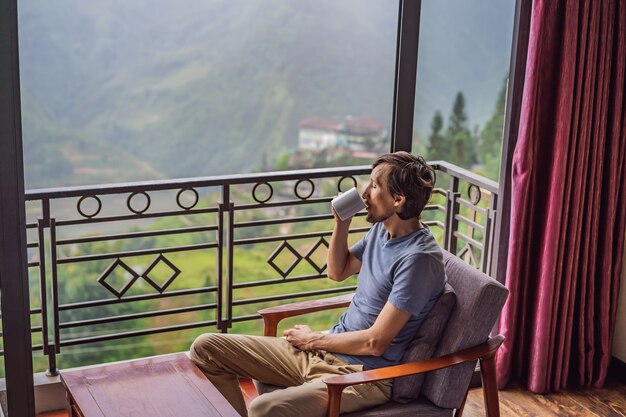 A young man holding coffee cup while sittinhg on chair on balcony looking at mountains and green