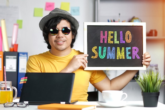 Young Man Holding Colorful Hello Summer Text on Blackboard While Working 