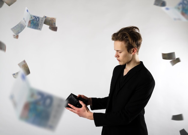 young man holding empty wallet by paper currency flying against white background