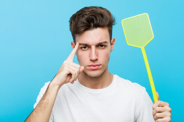 Young man holding a fly swatter pointing his temple with finger, thinking, focused on a task