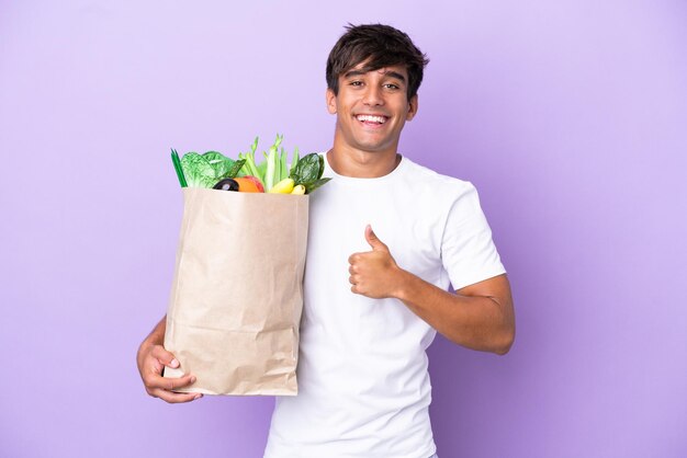 Молодой человек, держащий продуктовую сумку на фиолетовом фоне, показывает палец вверх