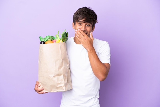 紫色の背景に分離された食料品の買い物袋を持っている若い男は、手で口を覆う幸せと笑顔