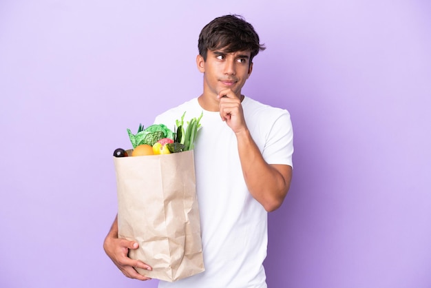 Young man holding a grocery shopping bag isolated on purple background having doubts and thinking