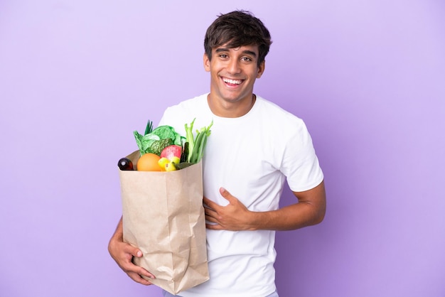 Young man holding a grocery shopping bag isolated on purple background smiling a lot