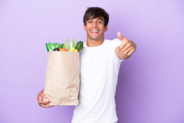 Young man holding a grocery shopping bag isolated on purple background with thumbs up because something good has happened