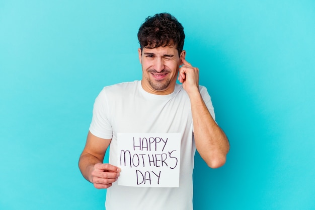 Young man holding a happy mothers day placard isolated covering ears with hands