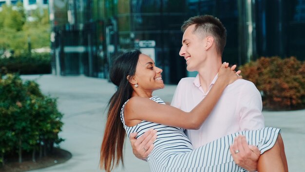 Young man holding his sweetheart in his hands and spinning around outdoors they have date in one sum...