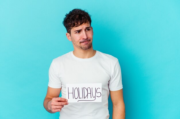 Young man holding a holidays placard isolated confused, feels doubtful and unsure