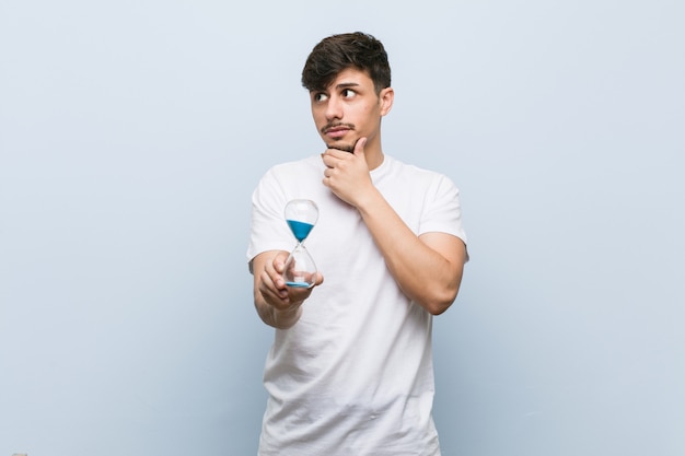 Young man holding a hourglass looking sideways with doubtful and skeptical expression