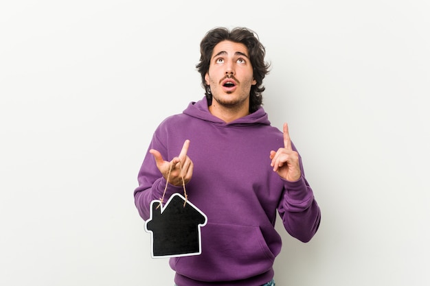 Young man holding a house icon shape pointing upside with opened mouth.