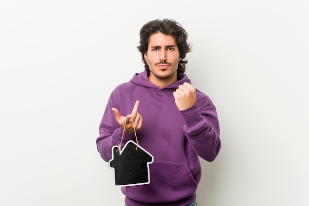 Молодой человек держа форму значка дома показывая кулак к камере, агрессивное выражение лица.