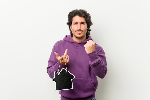 Young man holding a house icon shape showing fist to camera, aggressive facial expression.