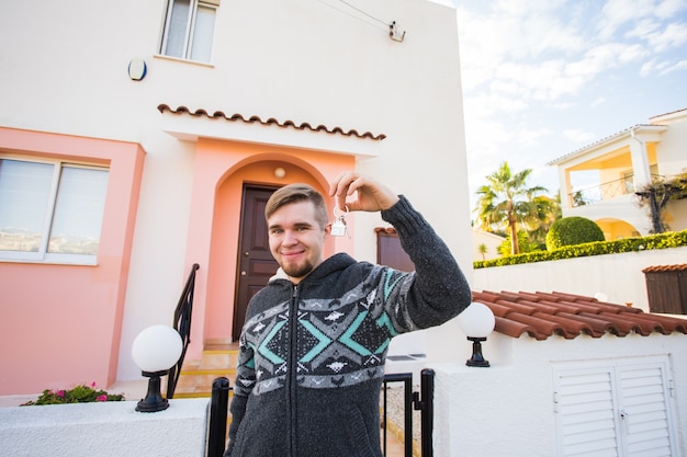 Young man holding house keys on house shaped keychain in front of a new home