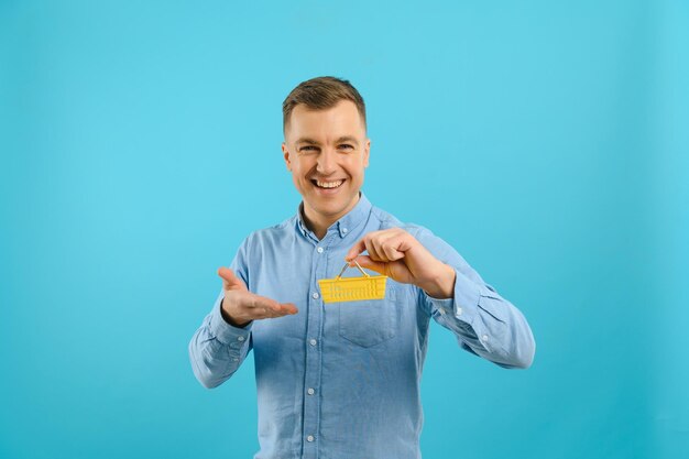 A young man holding a mini shopping cart and standing in front of a blue background in the studio Sales concept Place for text