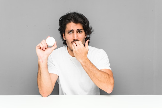 Young man holding a moisturizer biting fingernails, nervous and very anxious.