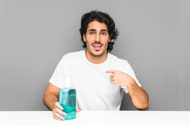 Young man holding a mouthwash surprised pointing at himself, smiling broadly.