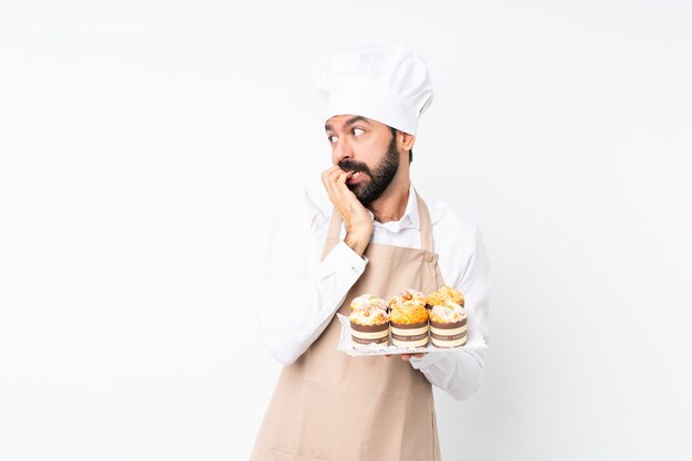 Young man holding muffin cake over isolated white wall nervous and scared putting hands to mouth