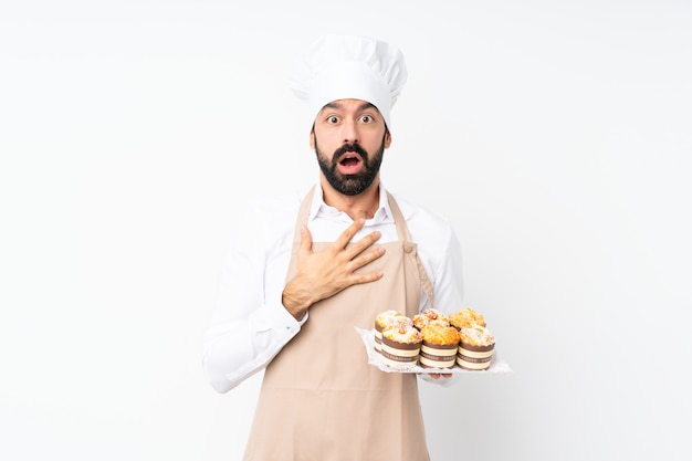 Young man holding muffin cake surprised and shocked while looking right