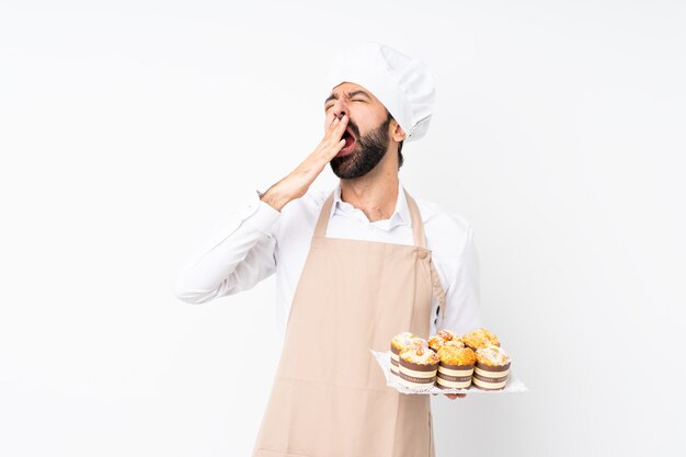 Young man holding muffin cake yawning and covering wide open mouth with hand