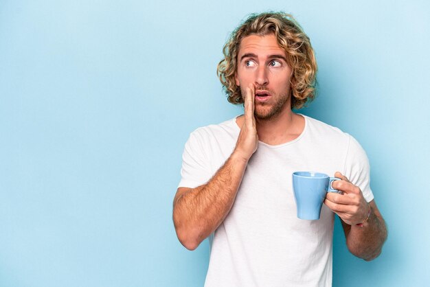 Young man holding a mug isolated on blue background is saying a secret hot braking news and looking aside