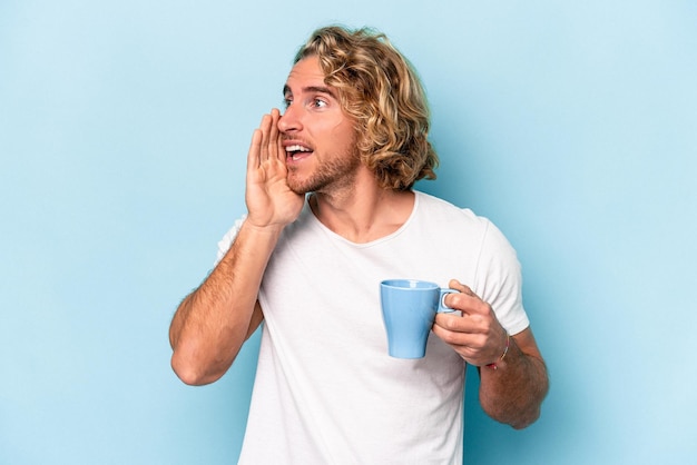 Young man holding a mug isolated on blue background shouting and holding palm near opened mouth.