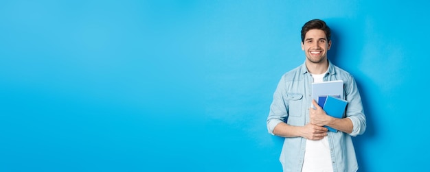 Young man holding notebooks and study material smiling happy standing over blue background