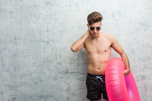 Photo young man holding a pink inflatable donut wearing a swimsuit suffering neck pain due to sedentary lifestyle.