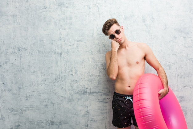 Young man holding a pink inflatable donut wearing a swimsuit who feels sad and pensive, looking at copy space.