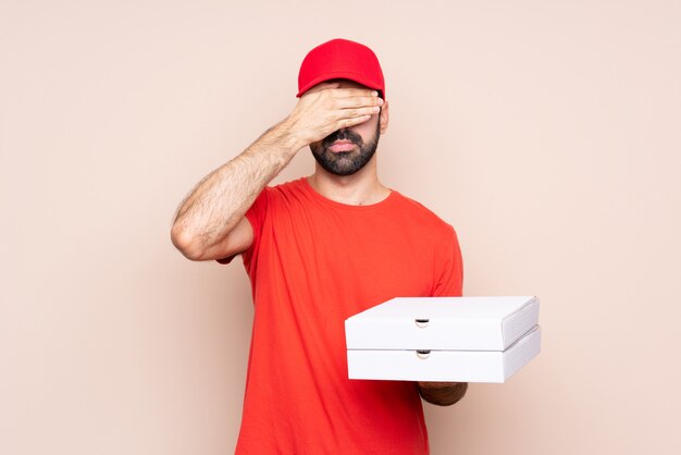Young man holding a pizza over isolated wall covering eyes by hands. Do not want to see something