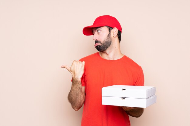 Young man holding a pizza over isolated wall unhappy and pointing to the side