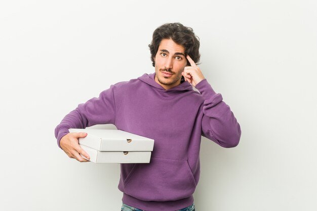 Young man holding pizzas package pointing his temple with finger, thinking, focused on a task