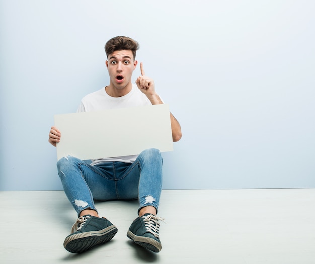 Young man holding a placard sitting on his home floor having some great idea, concept of creativity.