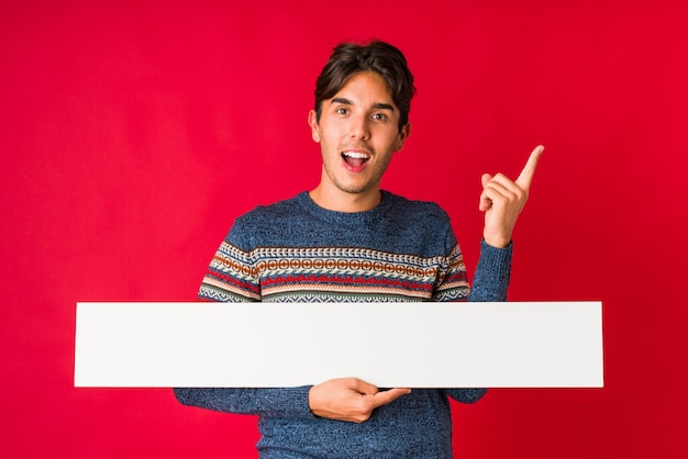 Young man holding a placard smiling cheerfully pointing with forefinger away.