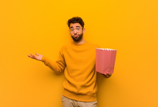 Young man holding popcorns doubting and shrugging shoulders