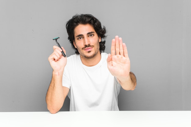 Young man holding a razor blade standing with outstretched hand showing stop sign, preventing you.