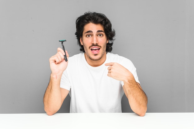 Young man holding a razor blade surprised pointing at himself, smiling broadly.