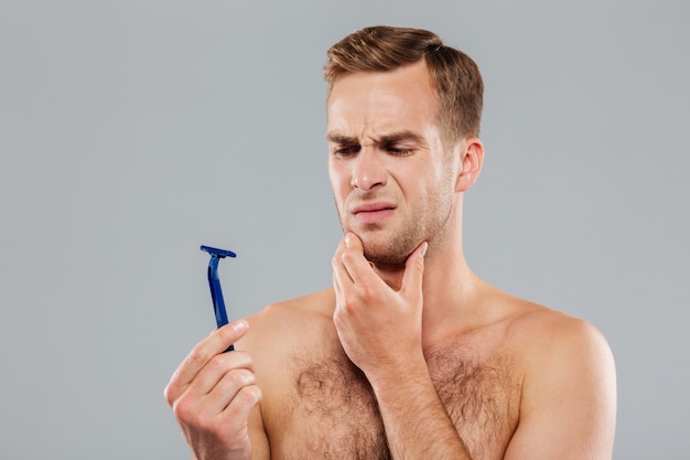 Photo young man holding razor and looking on it over gray wall