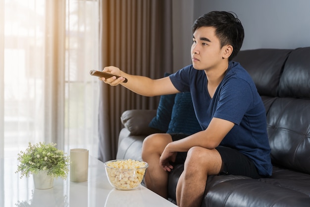 Young man holding remote control and watching TV while sitting on sofa 