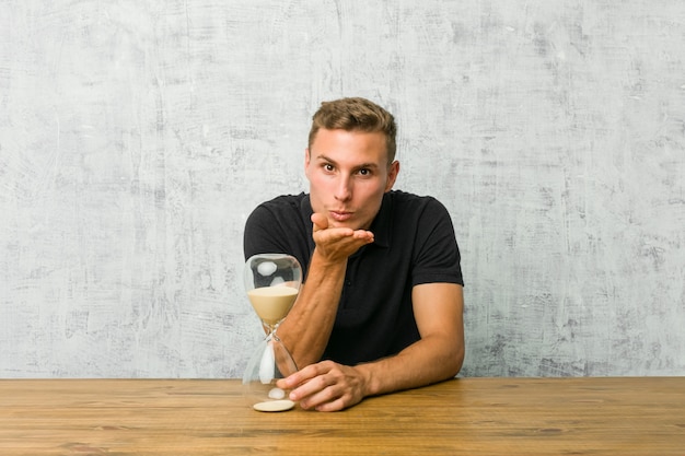 Young man holding a sand timer on a table folding lips and holding palms to send air kiss.